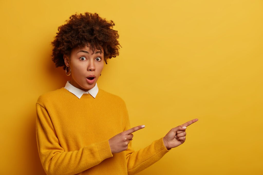 Amazed speechles young female model stares with shocked expression and points aside, tells about awesome discounts, wears casual yellow sweater, poses indoor, witensses something very astounding